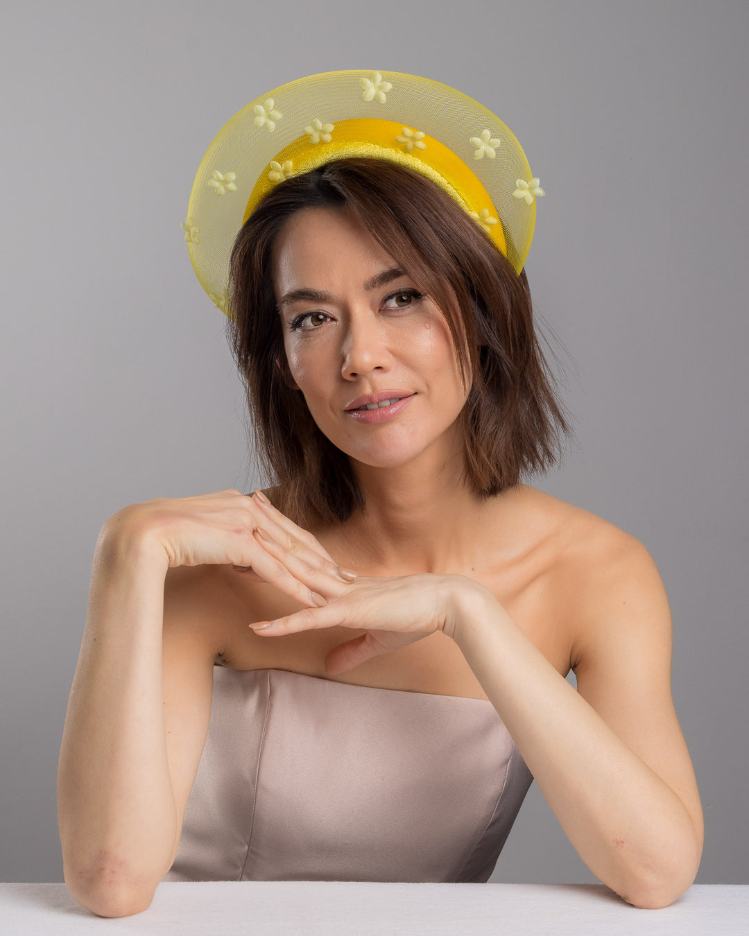 The female model is posing, wearing Soline, the lemon yellow headband fascinator featuring a satin-covered base encircled by a sheer structured halo, embellished with small tonal floral appliqués for a light, textured finish. Designed and made by Merve Bayindir Millinery.