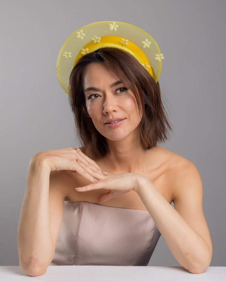 The female model is posing, wearing Soline, the lemon yellow headband fascinator featuring a satin-covered base encircled by a sheer structured halo, embellished with small tonal floral appliqués for a light, textured finish. Designed and made by Merve Bayindir Millinery.