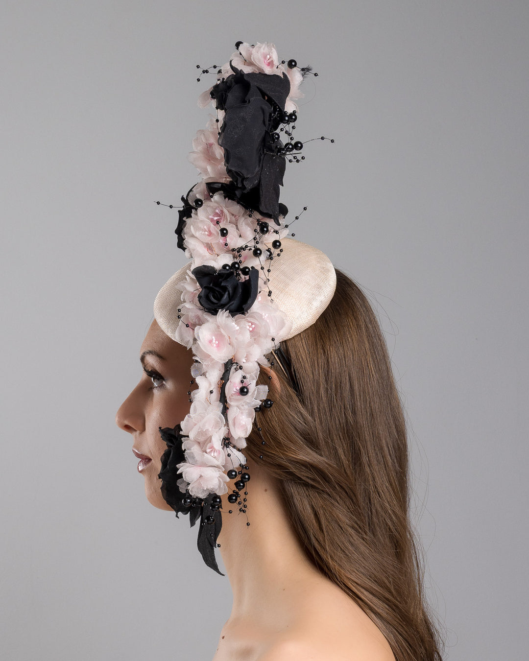 The model is showcasing the left side close up of the bush pink oval fascinator adorned with cascading black and soft pink chiffon flowers, accented with delicate black bead sprays, creating a dramatic, sculptural statement design. Designed and made by Merve Bayindir Millinery.