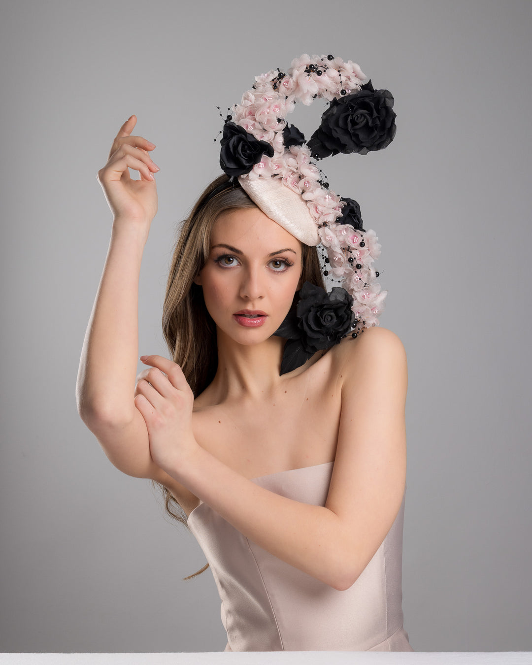 The female model is posing, wearing
Solstice, the bush pink oval fascinator adorned with cascading black and soft pink chiffon flowers, accented with delicate black bead sprays, creating a dramatic, sculptural statement design. Designed and made by Merve Bayindir Millinery.