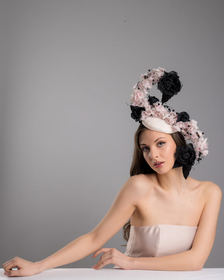 The female model is posing, wearing
Solstice, the bush pink oval fascinator adorned with cascading black and soft pink chiffon flowers, accented with delicate black bead sprays, creating a dramatic, sculptural statement design. Designed and made by Merve Bayindir Millinery.