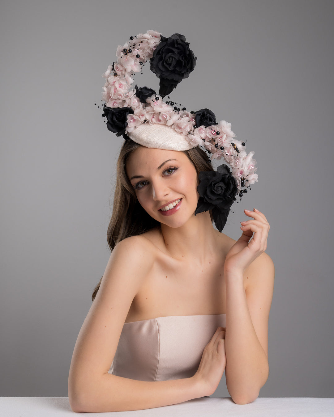 The female model is posing, wearing
Solstice, the bush pink oval fascinator adorned with cascading black and soft pink chiffon flowers, accented with delicate black bead sprays, creating a dramatic, sculptural statement design. Designed and made by Merve Bayindir Millinery.