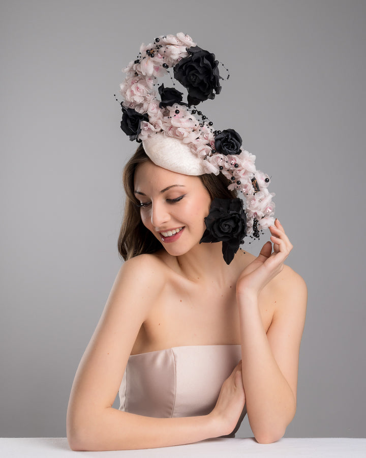 The female model is posing, wearing
Solstice, the bush pink oval fascinator adorned with cascading black and soft pink chiffon flowers, accented with delicate black bead sprays, creating a dramatic, sculptural statement design. Designed and made by Merve Bayindir Millinery.