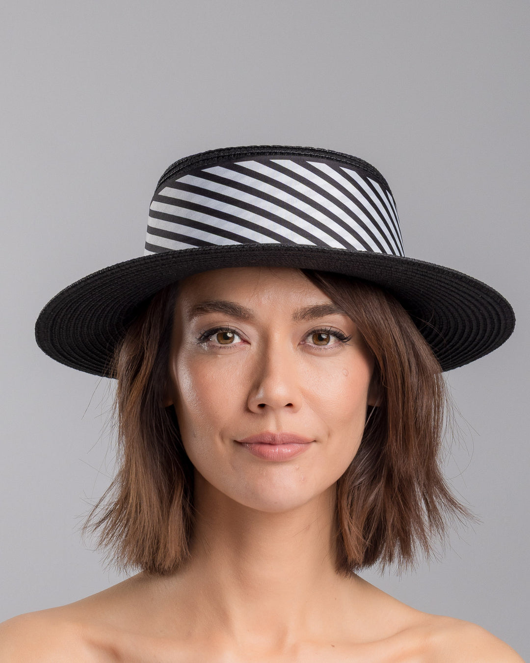 The model is showcasing the front side close up of the black woven straw boater hat with wide brim, finished with a black and white diagonal striped ribbon and a small red ladybug embellishment on the side. Designed and made by Merve Bayindir millinery.