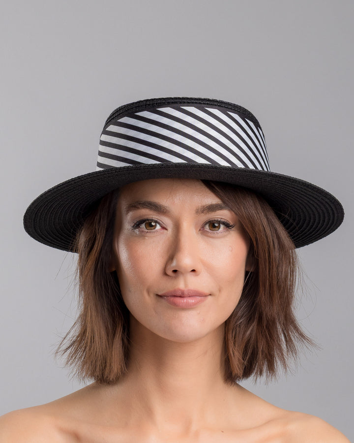 The model is showcasing the front side close up of the black woven straw boater hat with wide brim, finished with a black and white diagonal striped ribbon and a small red ladybug embellishment on the side. Designed and made by Merve Bayindir millinery.