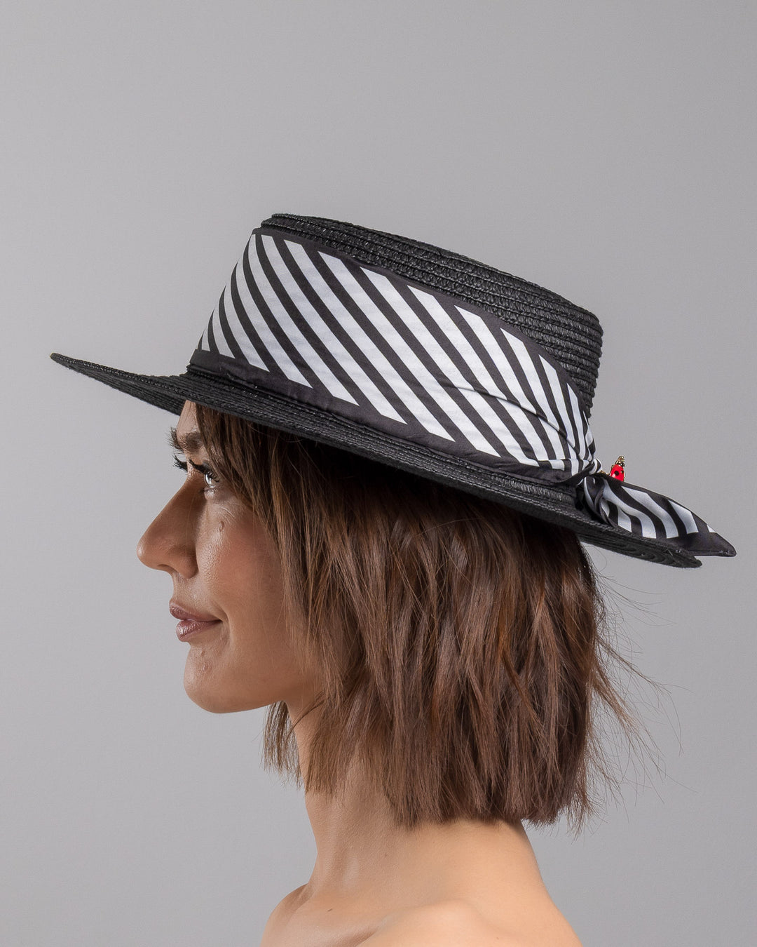 The model is showcasing the left side close up of the black woven straw boater hat with wide brim, finished with a black and white diagonal striped ribbon and a small red ladybug embellishment on the side. Designed and made by Merve Bayindir millinery.
