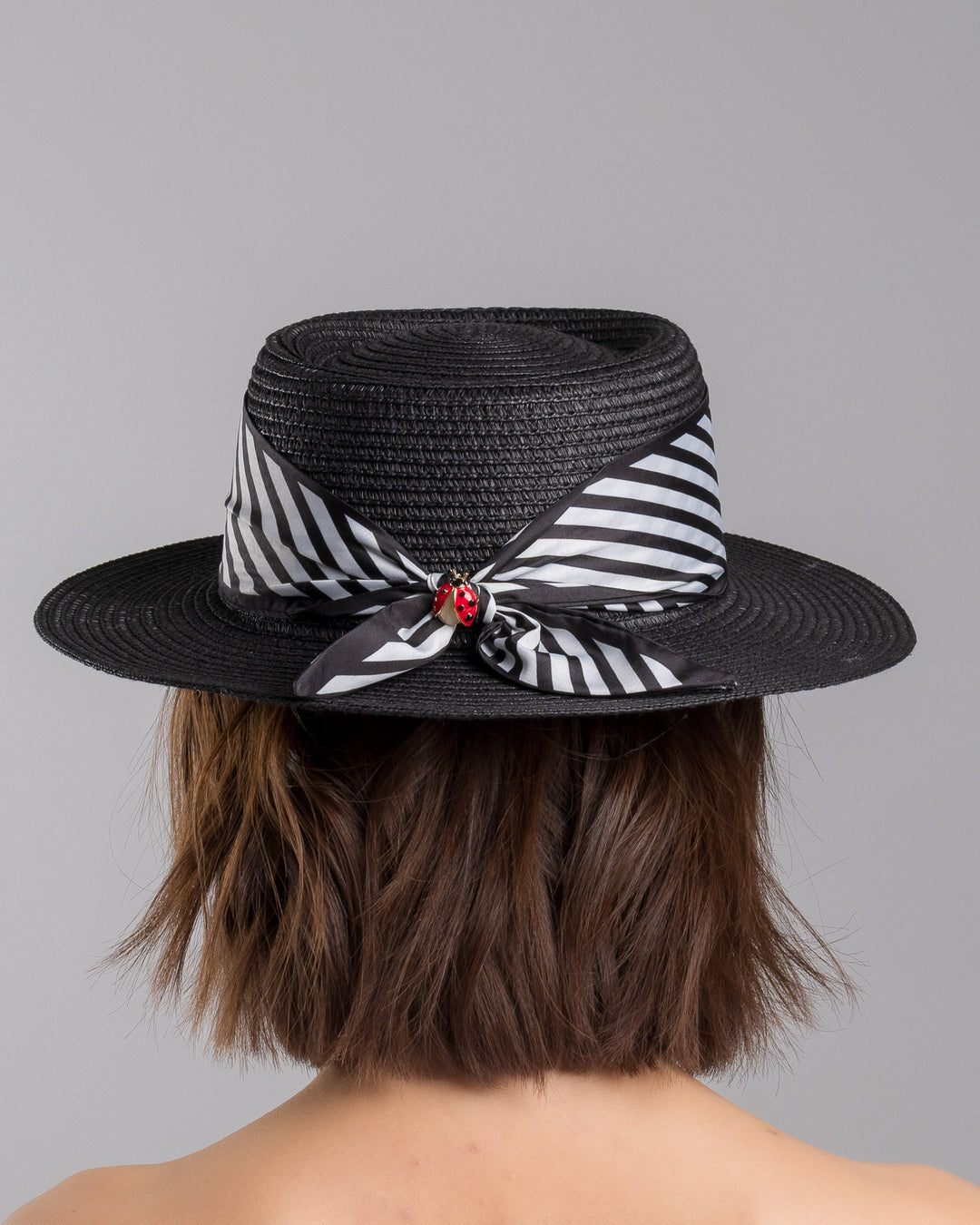 The model is showcasing the back side close up of the black woven straw boater hat with wide brim, finished with a black and white diagonal striped ribbon and a small red ladybug embellishment on the side. Designed and made by Merve Bayindir millinery.