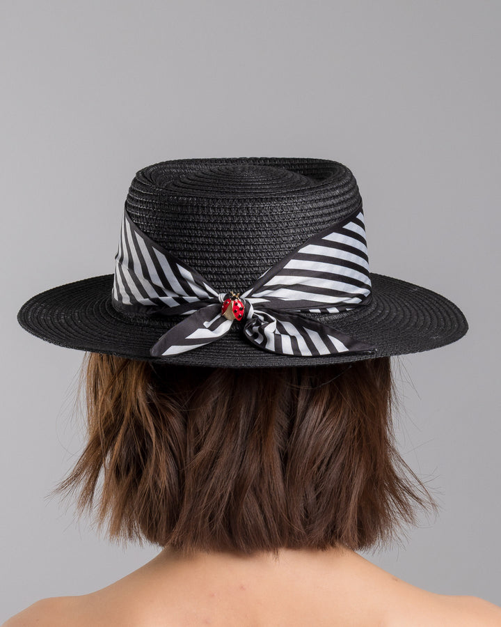 The model is showcasing the back side close up of the black woven straw boater hat with wide brim, finished with a black and white diagonal striped ribbon and a small red ladybug embellishment on the side. Designed and made by Merve Bayindir millinery.