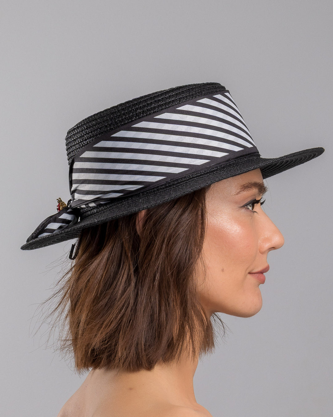The model is showcasing the right side close up of the black woven straw boater hat with wide brim, finished with a black and white diagonal striped ribbon and a small red ladybug embellishment on the side. Designed and made by Merve Bayindir millinery.