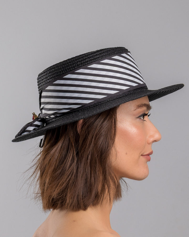 The model is showcasing the right side close up of the black woven straw boater hat with wide brim, finished with a black and white diagonal striped ribbon and a small red ladybug embellishment on the side. Designed and made by Merve Bayindir millinery.