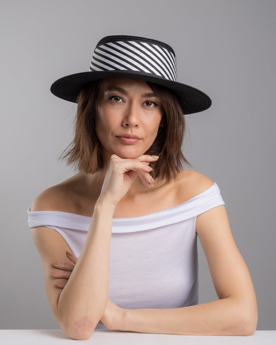 The female model is posing, wearing Stren, the black woven straw boater hat with wide brim, finished with a black and white diagonal striped ribbon and a small red ladybug embellishment on the side. Designed and made by Merve Bayindir millinery.