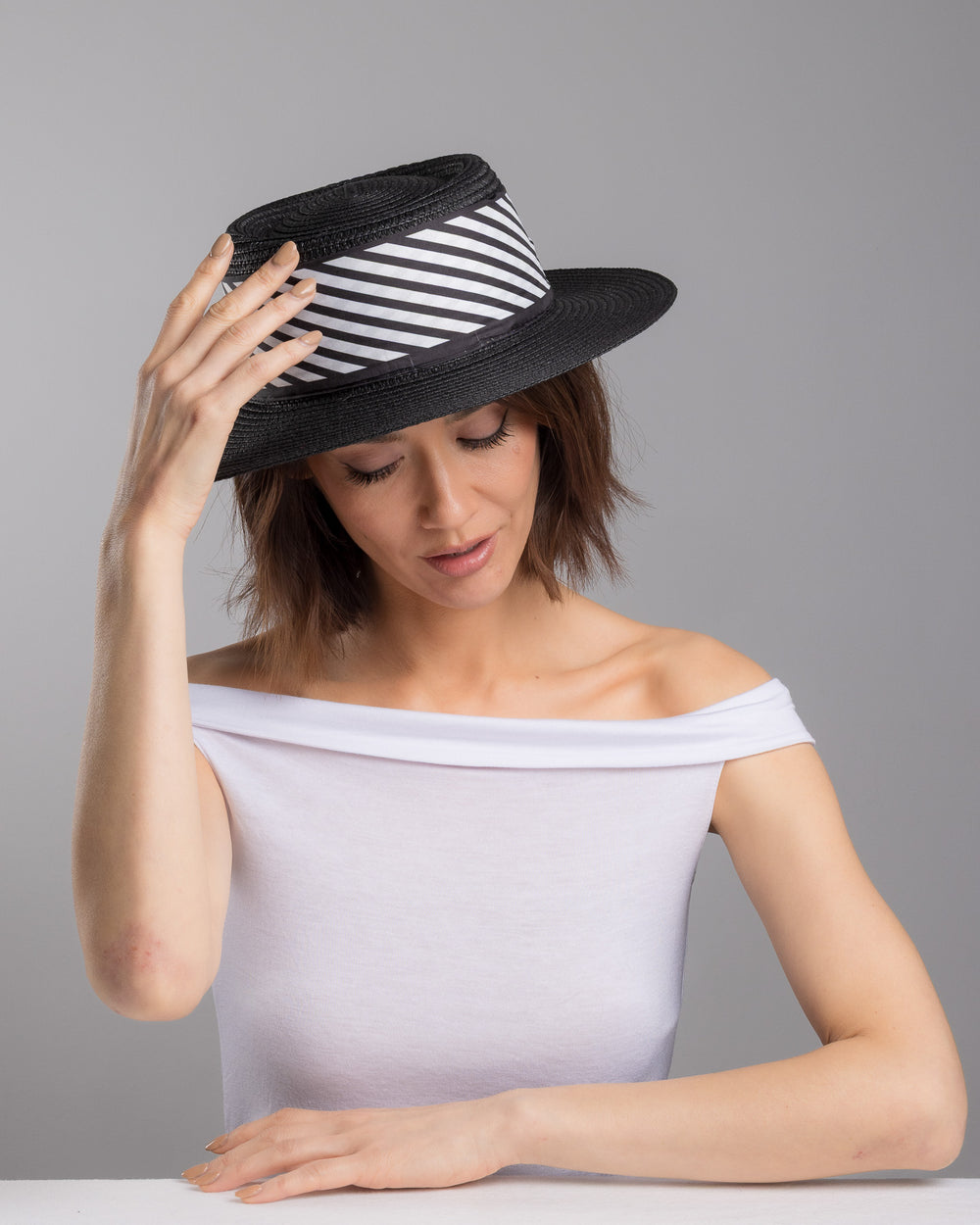 The female model is posing, wearing Stren, the black woven straw boater hat with wide brim, finished with a black and white diagonal striped ribbon and a small red ladybug embellishment on the side. Designed and made by Merve Bayindir millinery.