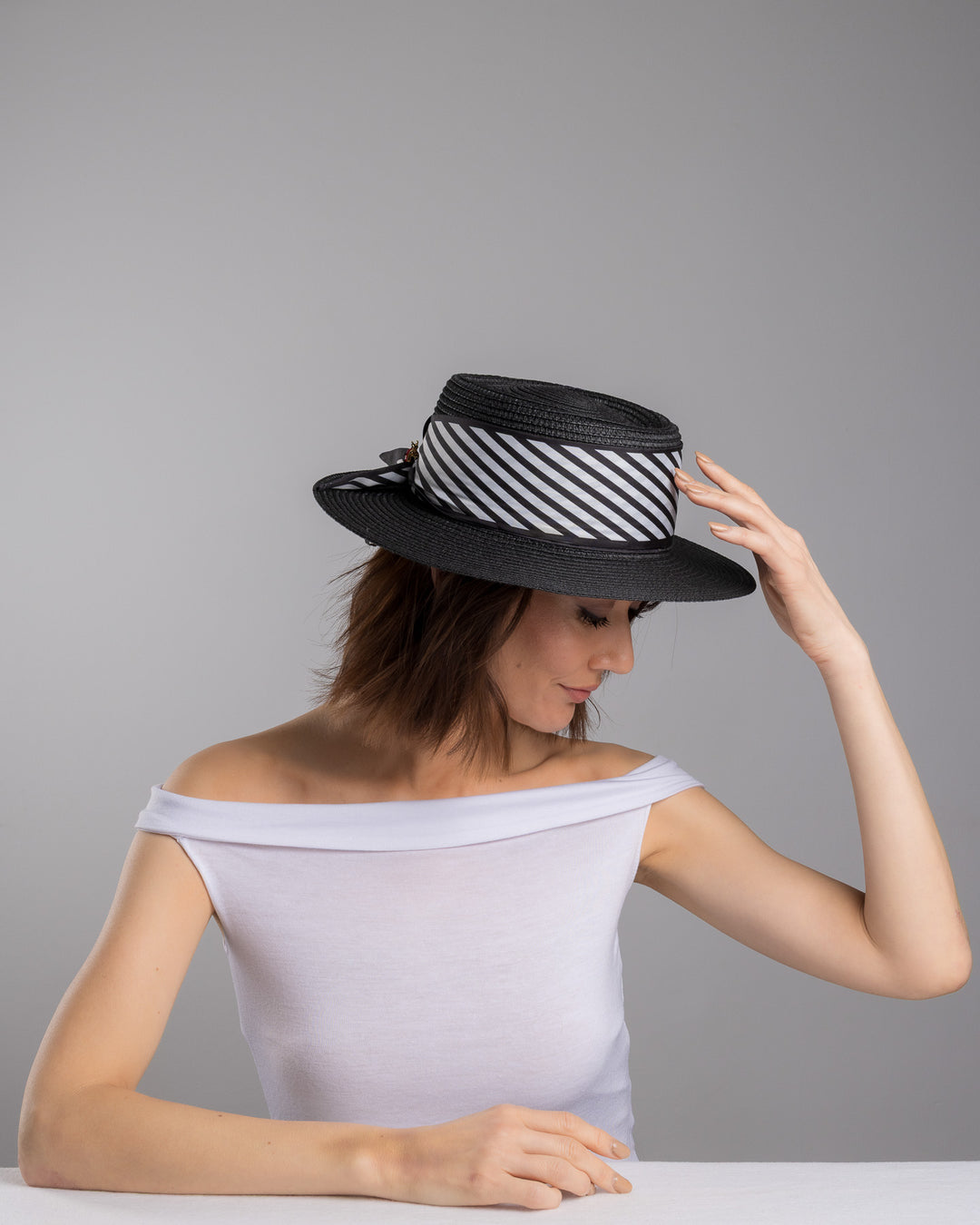 The female model is posing, wearing Stren, the black woven straw boater hat with wide brim, finished with a black and white diagonal striped ribbon and a small red ladybug embellishment on the side. Designed and made by Merve Bayindir millinery.