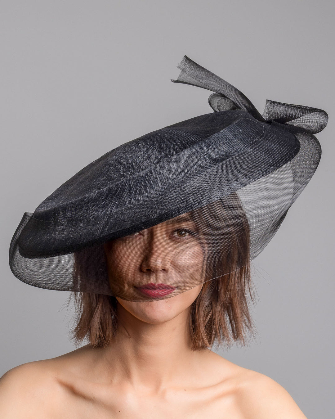 The model is showcasing the front side close up of the black structured pillbox hat crafted from a grade sinamay, featuring a soft sheer veil overlay that drapes around the base and a delicate bow detail at the back. Designed and made by Merve Bayindir Millinery.
