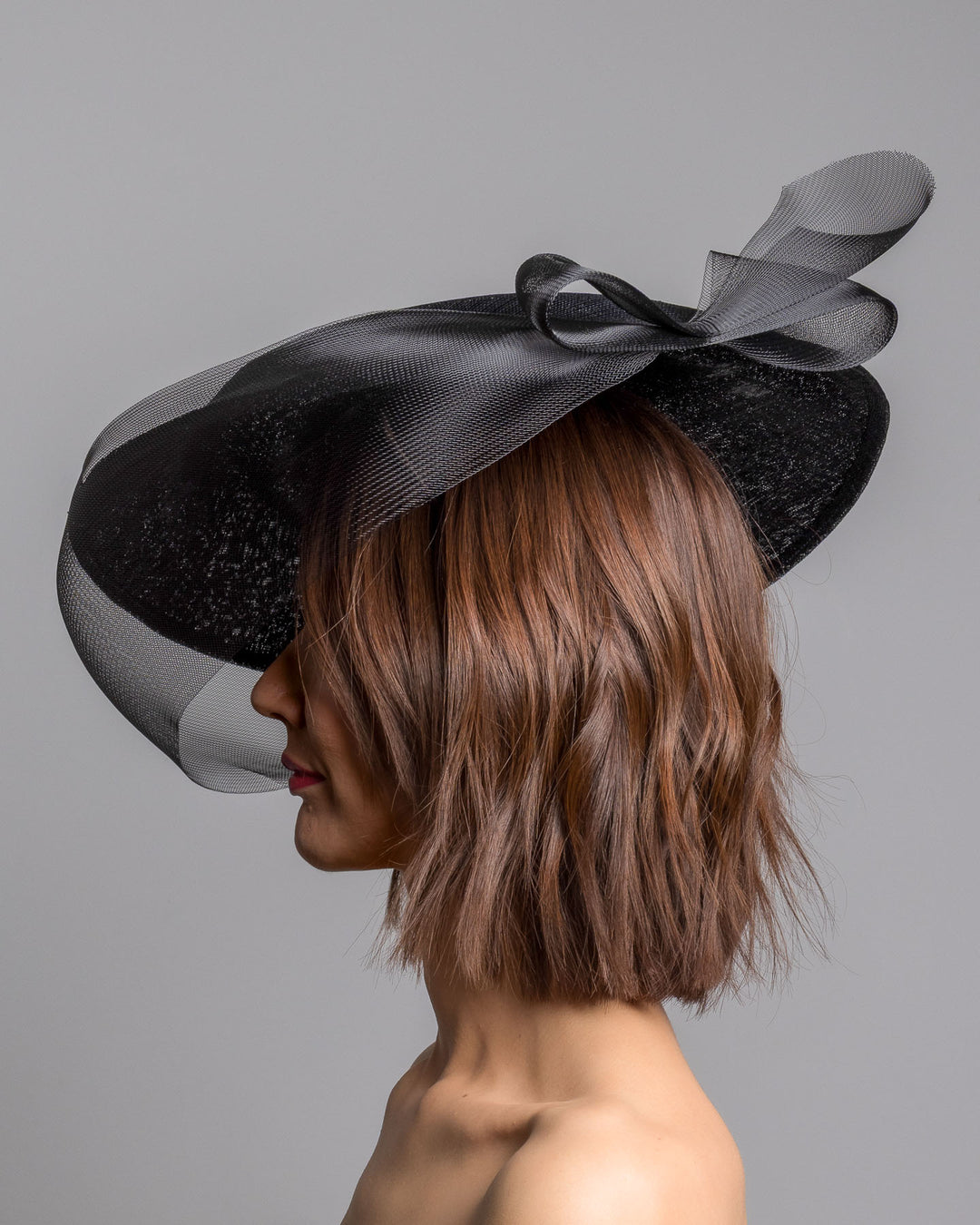 The model is showcasing the left side close up of the black structured pillbox hat crafted from a grade sinamay, featuring a soft sheer veil overlay that drapes around the base and a delicate bow detail at the back. Designed and made by Merve Bayindir Millinery.
