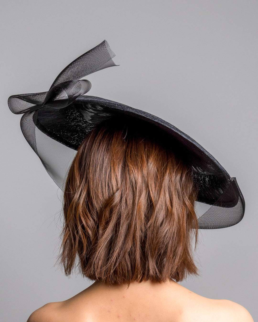 The model is showcasing the back side close up of the black structured pillbox hat crafted from a grade sinamay, featuring a soft sheer veil overlay that drapes around the base and a delicate bow detail at the back. Designed and made by Merve Bayindir Millinery.
