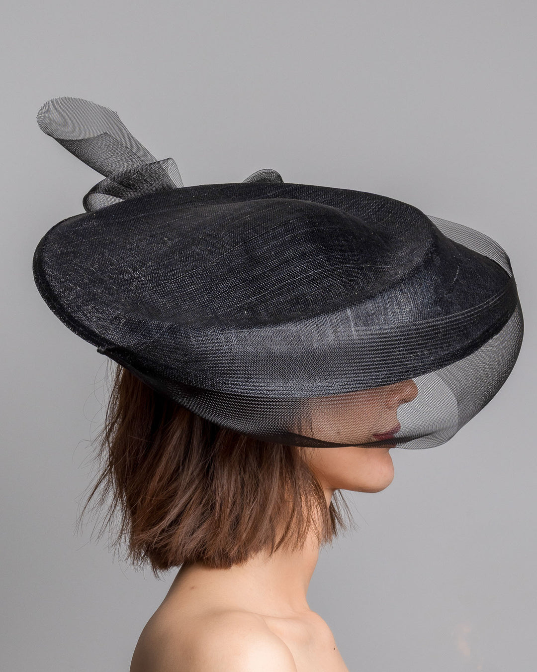 The model is showcasing the right side close up of the black structured pillbox hat crafted from a grade sinamay, featuring a soft sheer veil overlay that drapes around the base and a delicate bow detail at the back. Designed and made by Merve Bayindir Millinery.
