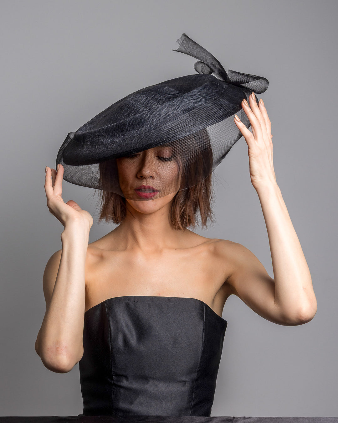 The female model is posing, wearing 
Sveil, the black structured pillbox hat crafted from a grade sinamay, featuring a soft sheer veil overlay that drapes around the base and a delicate bow detail at the back. Designed and made by Merve Bayindir Millinery.
