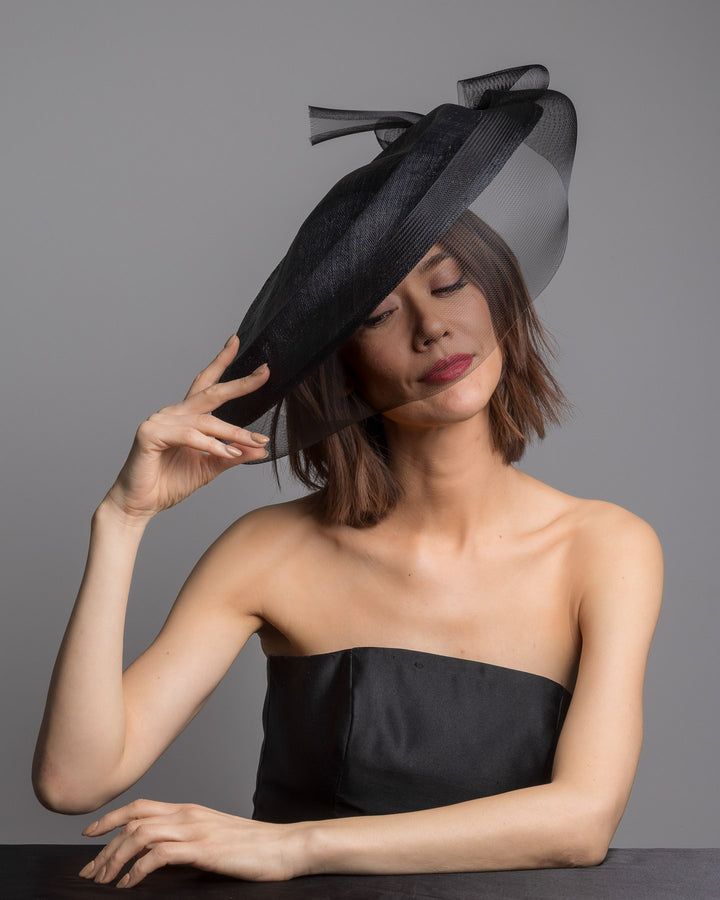 The female model is posing, wearing 
Sveil, the black structured pillbox hat crafted from a grade sinamay, featuring a soft sheer veil overlay that drapes around the base and a delicate bow detail at the back. Designed and made by Merve Bayindir Millinery.
