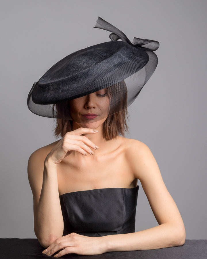 The female model is posing, wearing 
Sveil, the black structured pillbox hat crafted from a grade sinamay, featuring a soft sheer veil overlay that drapes around the base and a delicate bow detail at the back. Designed and made by Merve Bayindir Millinery.
