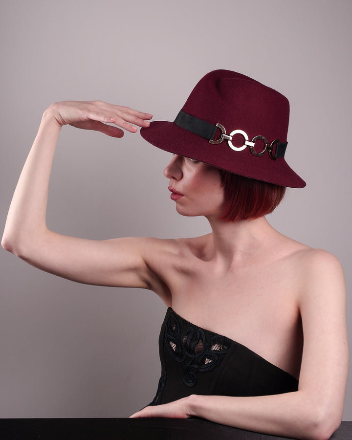 The model showcases the left front from aboce angle of the Sable Fedora hat, which is crafted from rich burgundy felt and finished with a sleek black band and bold gold-coloured metal chain detailing. Designed by Merve Bayindir.
