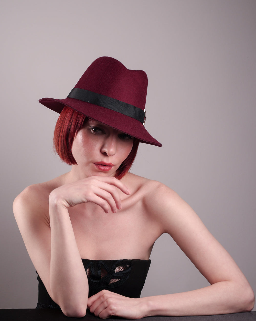 The model showcases the front of the Sable Fedora hat, which is crafted from rich burgundy felt and finished with a sleek black band and bold gold-coloured metal chain detailing. Designed by Merve Bayindir.