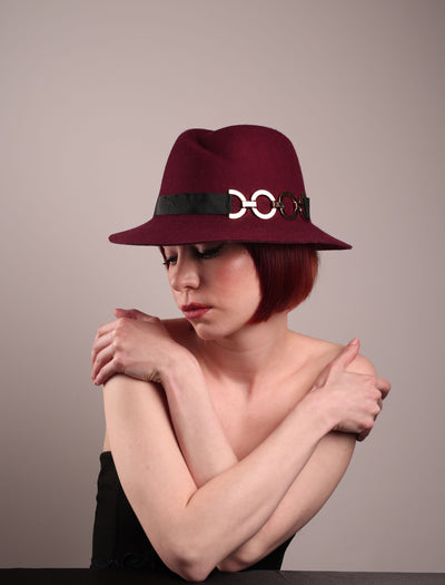 The model showcases the left front side of the Sable Fedora hat, which is crafted from rich burgundy felt and finished with a sleek black band and bold gold-coloured metal chain detailing. Designed by Merve Bayindir.