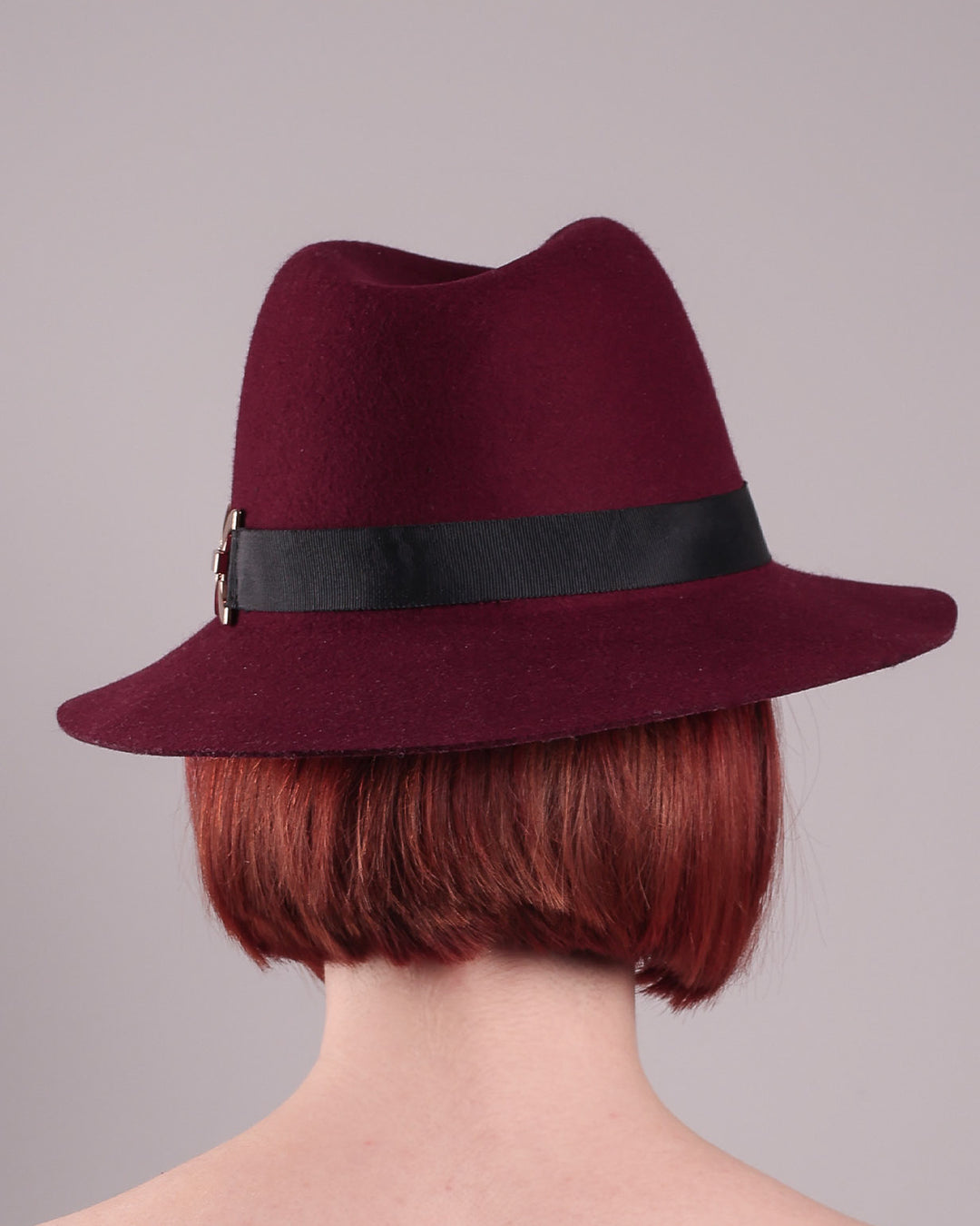 The model showcases the back of the Sable Fedora hat, which is crafted from rich burgundy felt and finished with a sleek black band and bold gold-coloured metal chain detailing. Designed by Merve Bayindir.