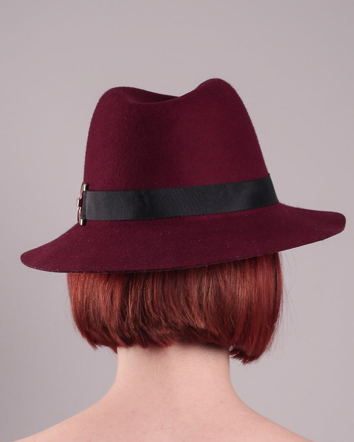 The model showcases the back of the Sable Fedora hat, which is crafted from rich burgundy felt and finished with a sleek black band and bold gold-coloured metal chain detailing. Designed by Merve Bayindir.