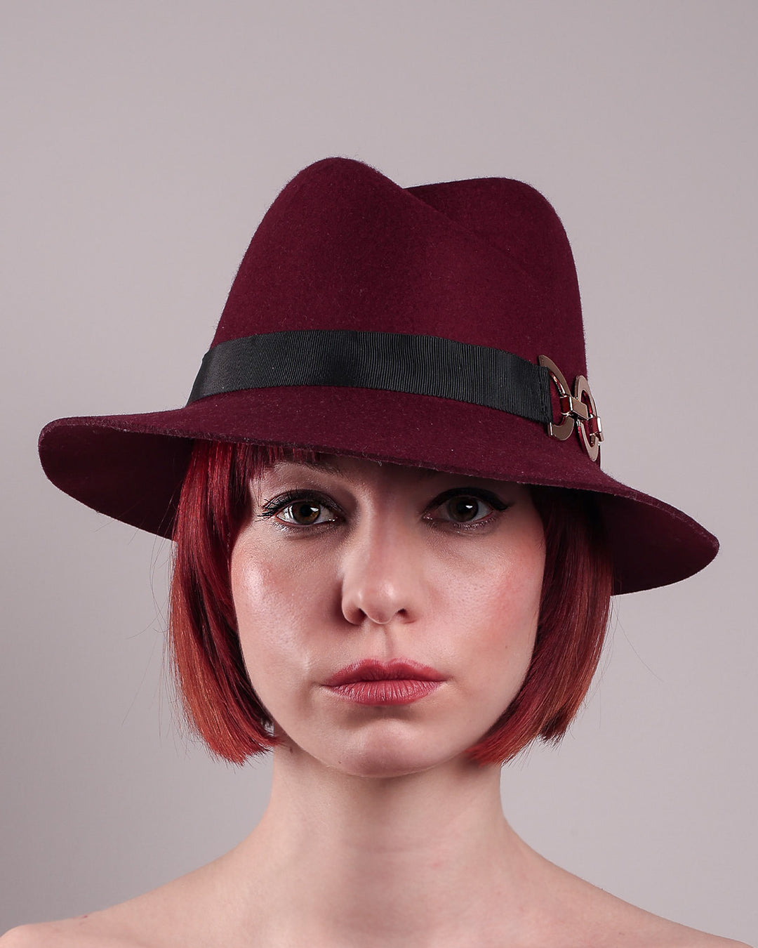 The model showcases the front close up of the Sable Fedora hat, which is crafted from rich burgundy felt and finished with a sleek black band and bold gold-coloured metal chain detailing. Designed by Merve Bayindir.
