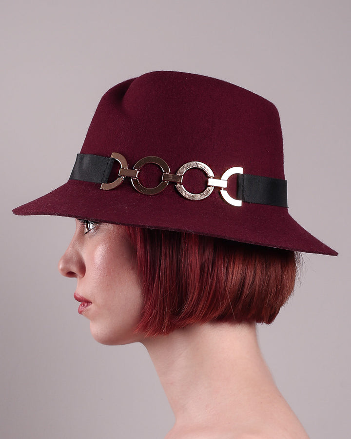 The model showcases the front close up of the Sable Fedora hat, which is crafted from rich burgundy felt and finished with a sleek black band and bold gold-coloured metal chain detailing. Designed by Merve Bayindir.