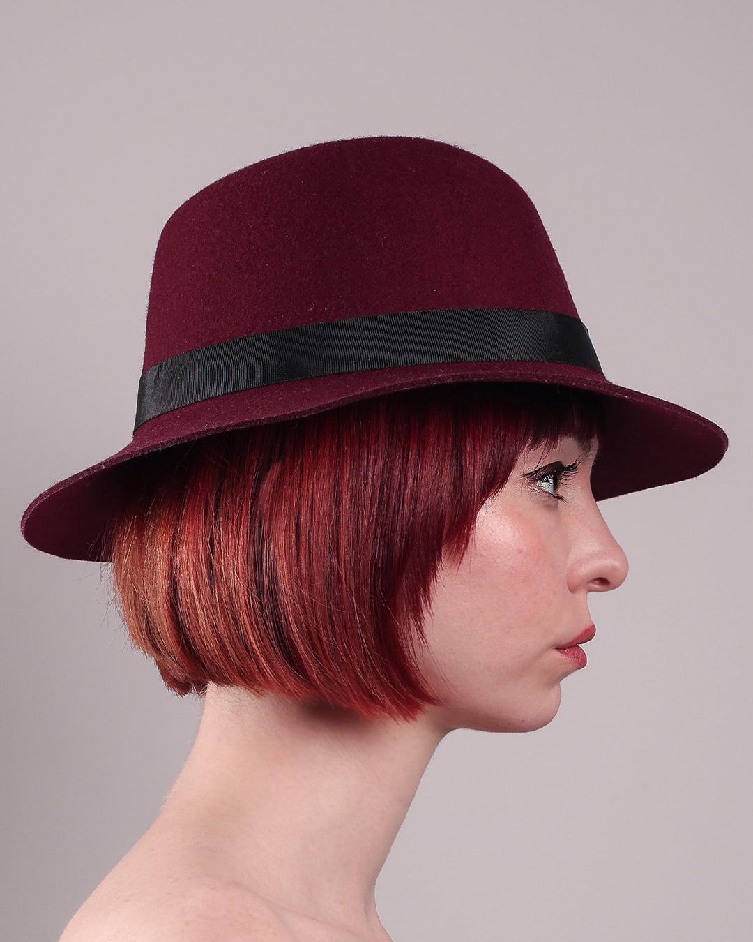 The model showcases the right side close up of the Sable Fedora hat, which is crafted from rich burgundy felt and finished with a sleek black band and bold gold-coloured metal chain detailing. Designed by Merve Bayindir.