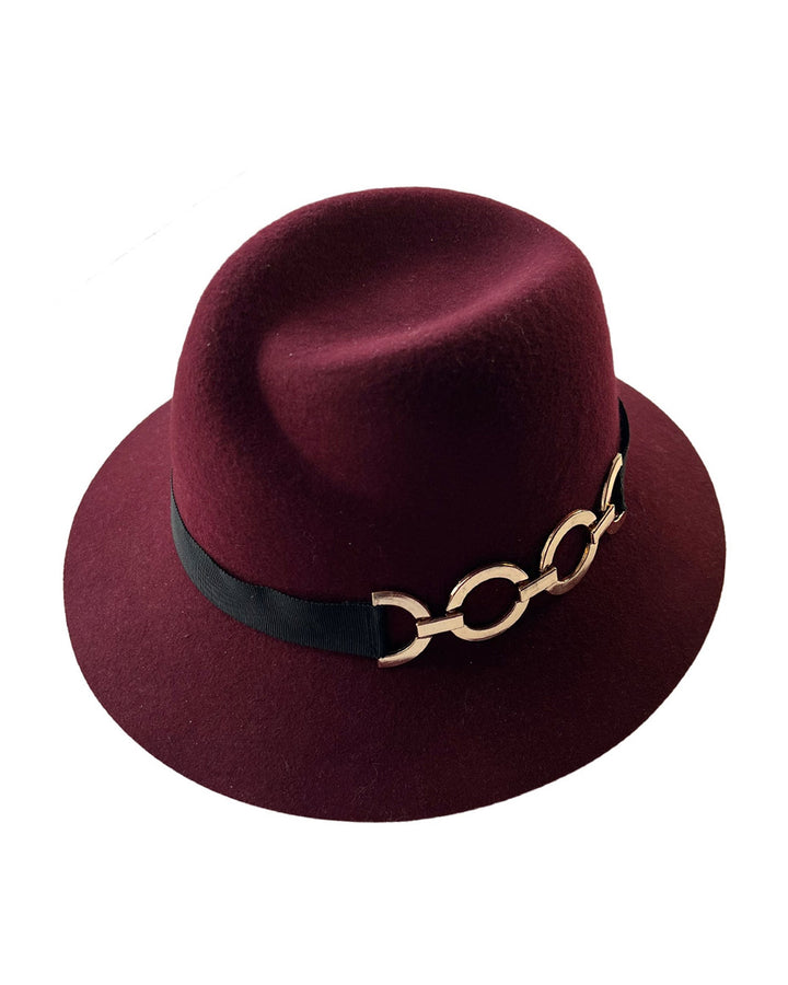 Still life picture of the Sable Fedora hat, which is crafted from rich burgundy felt and finished with a sleek black band and bold gold-coloured metal chain detailing. Designed by Merve Bayindir.