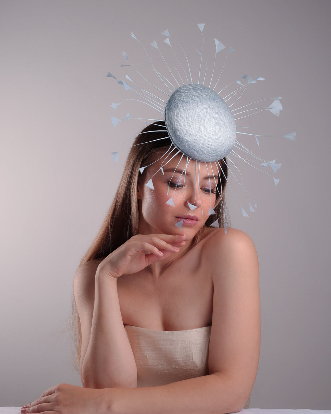 The model is showcasing the right front side of the double button light blue fascinator, decorated with blue goose feathers all around angle it like sun, fitted with a headband and elastic for secure fitting.