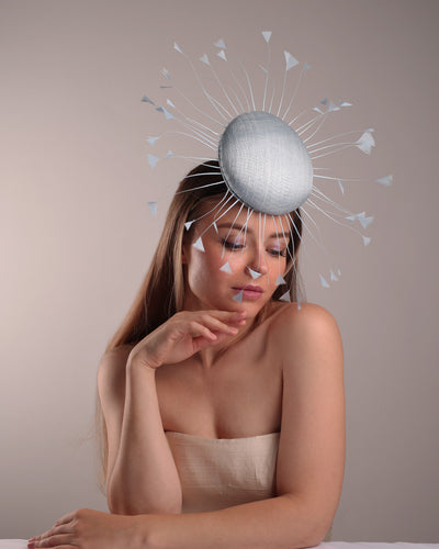 The model is showcasing the right front side of the double button light blue fascinator, decorated with blue goose feathers all around angle it like sun, fitted with a headband and elastic for secure fitting.