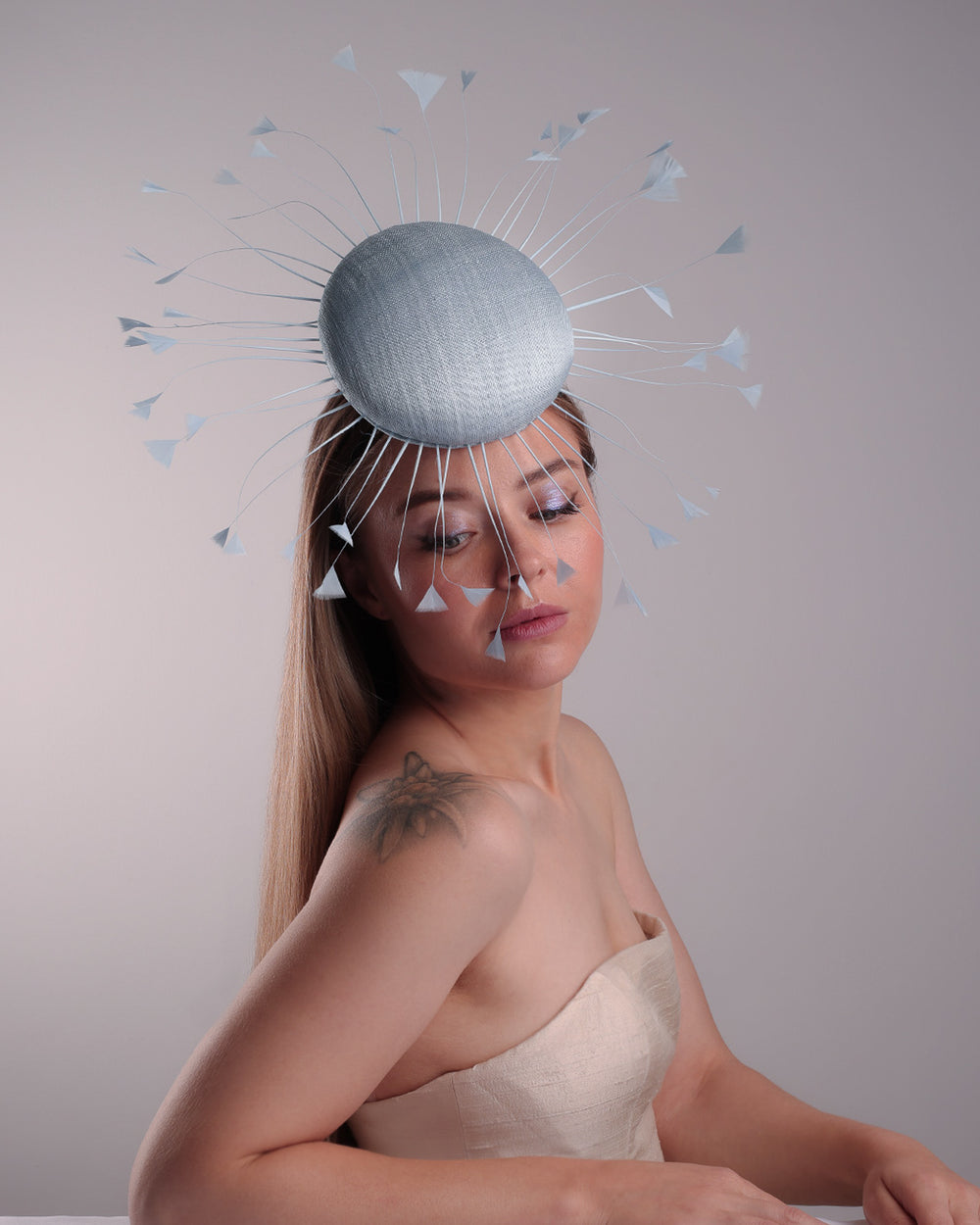 The model is showcasing the front side of the double button light blue fascinator, decorated with blue goose feathers all around angle it like sun, fitted with a headband and elastic for secure fitting.