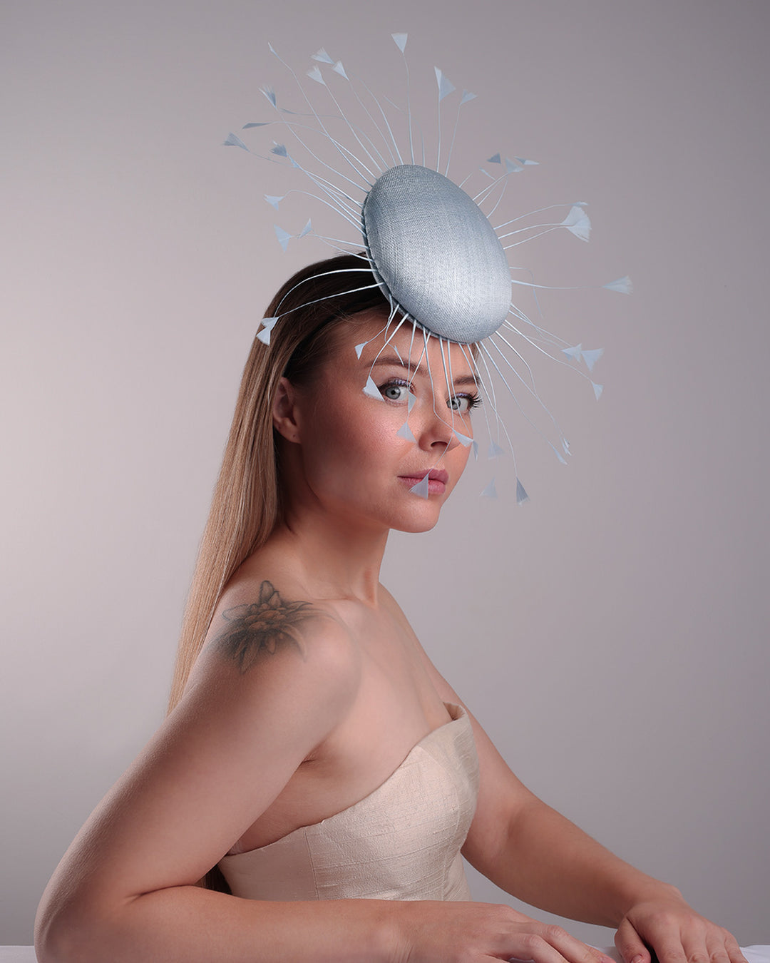 The model is showcasing the right front side angle of the double button light blue fascinator, decorated with blue goose feathers all around angle it like sun, fitted with a headband and elastic for secure fitting.