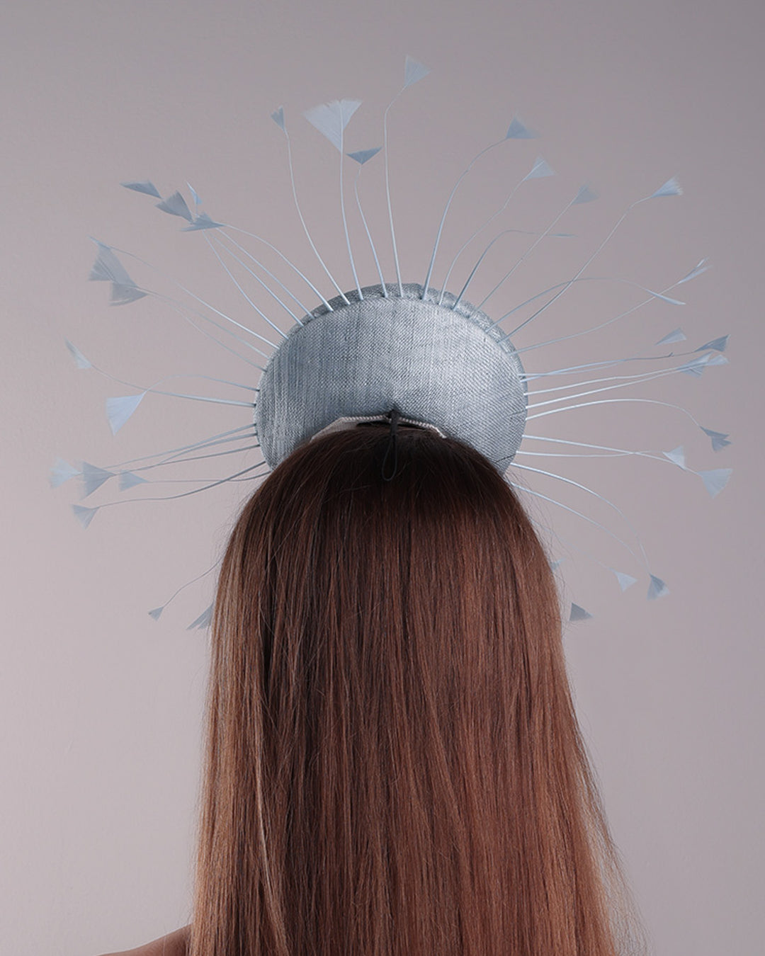 The model is showcasing the back of the double button light blue fascinator, decorated with blue goose feathers all around angle it like sun, fitted with a headband and elastic for secure fitting.