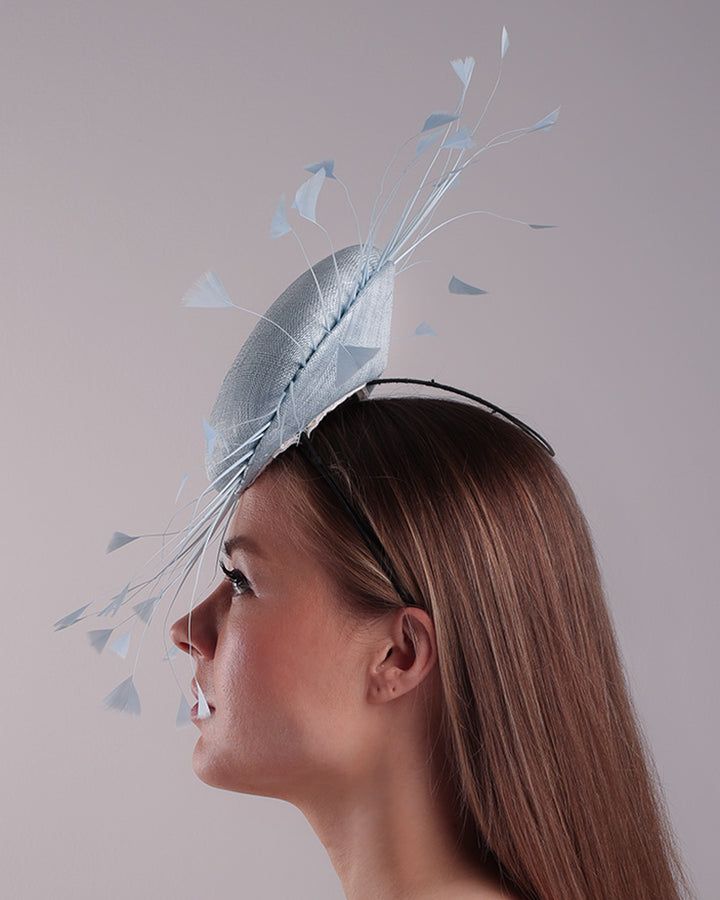 The model is showcasing the left side of the double button light blue fascinator, decorated with blue goose feathers all around angle it like sun, fitted with a headband and elastic for secure fitting.