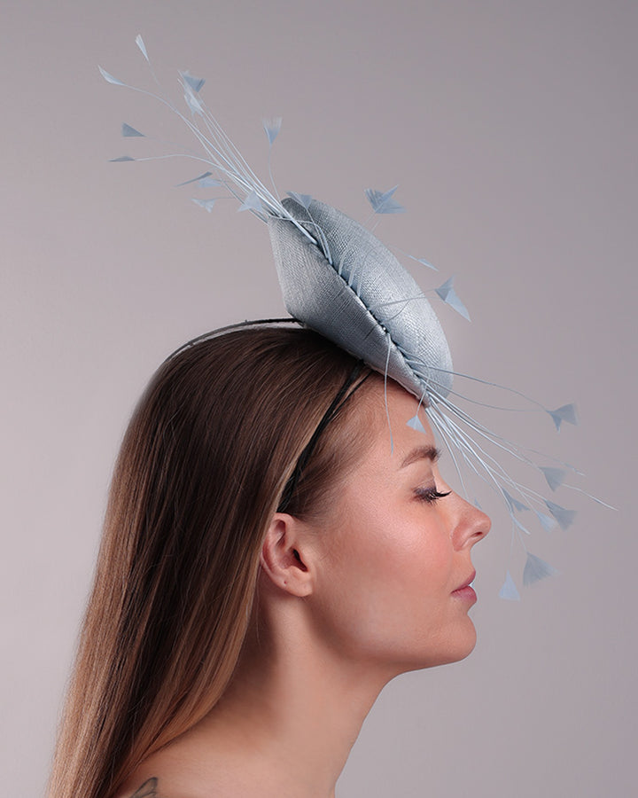 The model is showcasing the right side of the double button light blue fascinator, decorated with blue goose feathers all around angle it like sun, fitted with a headband and elastic for secure fitting.