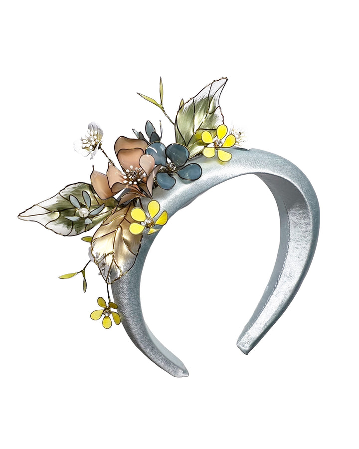 Powder blue satin padded headband featuring an asymmetrical arrangement of handcrafted translucent flowers and leaves in soft peach, dusty blue, pale yellow, and muted green tones. The petals are delicately edged with fine gold detailing and accented with subtle crystal centers and beadwork, creating an elegant, sculptural statement piece ideal for weddings and refined special occasions. Designed and made by Merve Bayindir Millinery.

