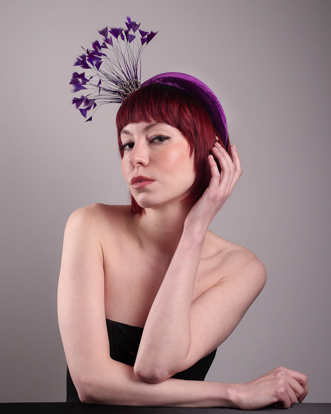 The model showcases the left front of the Seraphina headband fascinator, wrapped in plush violet velvet. This sculptural piece bursts into movement with a spray of deep purple feathers and a gold-coloured metal fan featuring branch detailing on the right front side. Designed by Merve Bayindir.