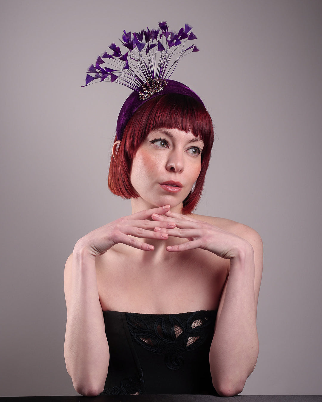 The model showcases the right front of the Seraphina headband fascinator, wrapped in plush violet velvet. This sculptural piece bursts into movement with a spray of deep purple feathers and a gold-coloured metal fan featuring branch detailing on the right front side. Designed by Merve Bayindir.