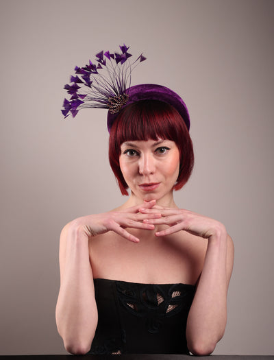 The model showcases the front of the Seraphina headband fascinator, wrapped in plush violet velvet. This sculptural piece bursts into movement with a spray of deep purple feathers and a gold-coloured metal fan featuring branch detailing on the right front side. Designed by Merve Bayindir.