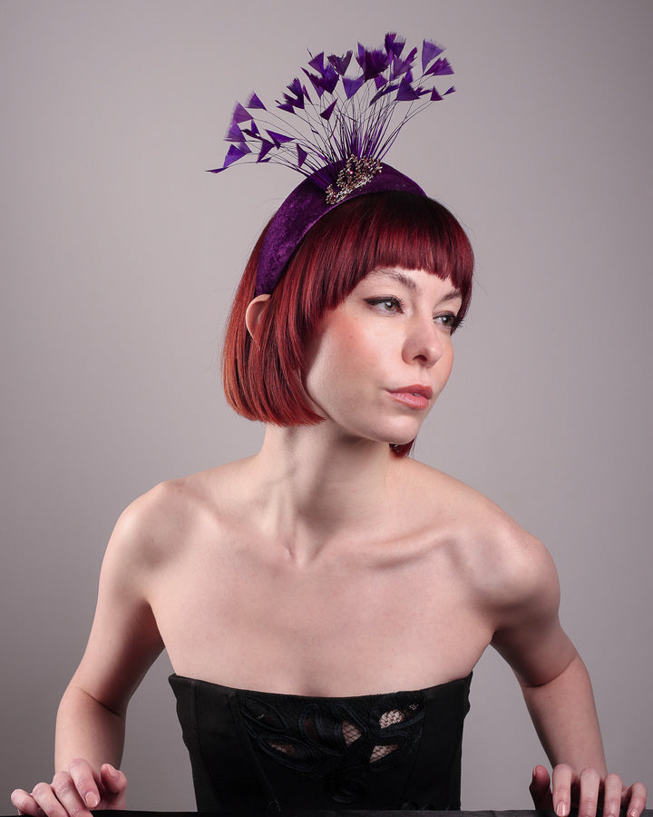 The model showcases the right side focused picture of the Seraphina headband fascinator, wrapped in plush violet velvet. This sculptural piece bursts into movement with a spray of deep purple feathers and a gold-coloured metal fan featuring branch detailing on the right front side. Designed by Merve Bayindir.
