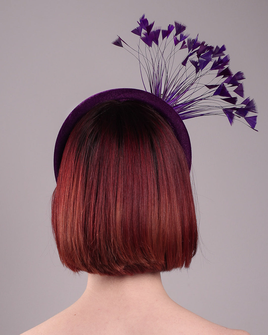 The model showcases the back of the Seraphina headband fascinator, wrapped in plush violet velvet. This sculptural piece bursts into movement with a spray of deep purple feathers and a gold-coloured metal fan featuring branch detailing on the right front side. Designed by Merve Bayindir.