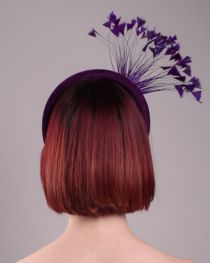The model showcases the back of the Seraphina headband fascinator, wrapped in plush violet velvet. This sculptural piece bursts into movement with a spray of deep purple feathers and a gold-coloured metal fan featuring branch detailing on the right front side. Designed by Merve Bayindir.