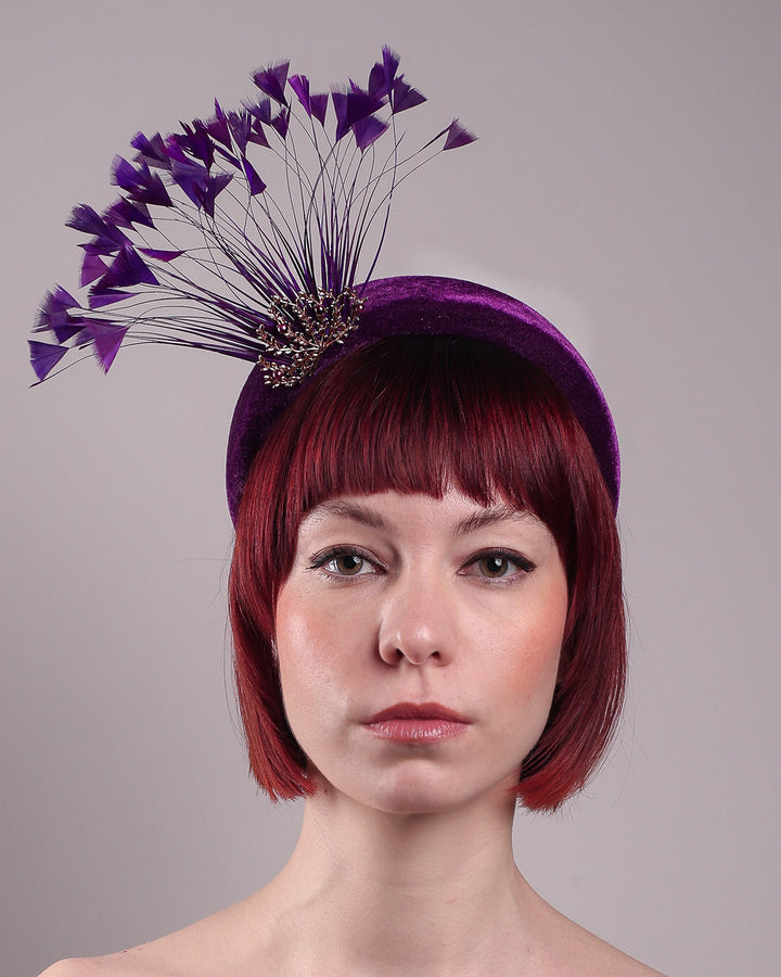 The model showcases the front close up of the Seraphina headband fascinator, wrapped in plush violet velvet. This sculptural piece bursts into movement with a spray of deep purple feathers and a gold-coloured metal fan featuring branch detailing on the right front side. Designed by Merve Bayindir.