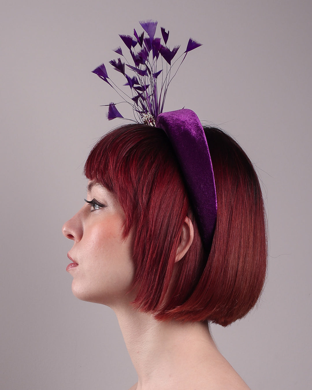 The model showcases the left side close up of the Seraphina headband fascinator, wrapped in plush violet velvet. This sculptural piece bursts into movement with a spray of deep purple feathers and a gold-coloured metal fan featuring branch detailing on the right front side. Designed by Merve Bayindir.
