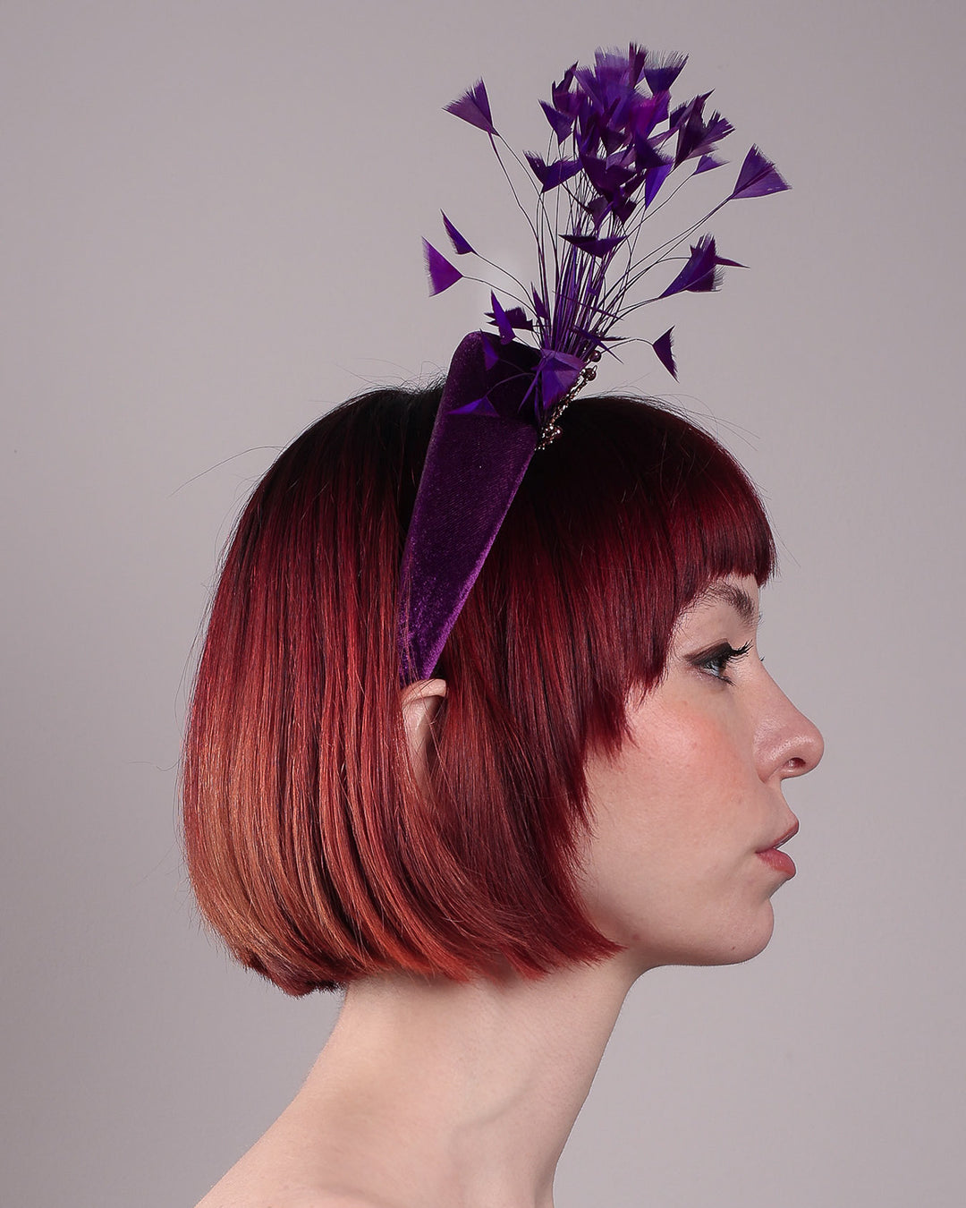 The model showcases the back of the Seraphina headband fascinator, wrapped in plush violet velvet. This sculptural piece bursts into movement with a spray of deep purple feathers and a gold-coloured metal fan featuring branch detailing on the right front side. Designed by Merve Bayindir.